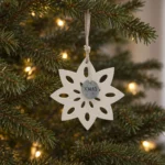 Vit snöflinga i trä med ovala utskärningar och en rund XMAS-bricka, hängande i en julgran med varm belysning.