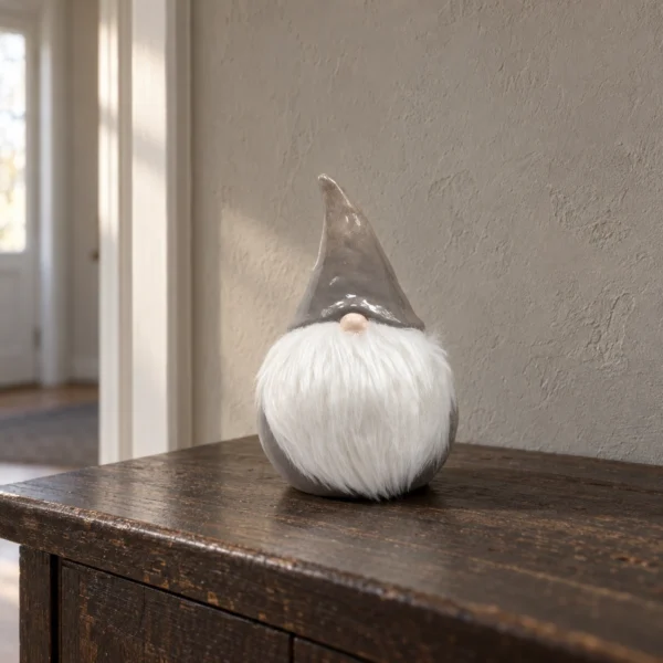 Tomte med spetsig mössa och vitt textilskägg på en mörk träyta framför en varmgrå vägg
