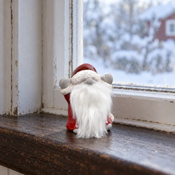 Liten tomte med röd luva, grå vantar och vitt skägg på en fönsterbräda i trä.
