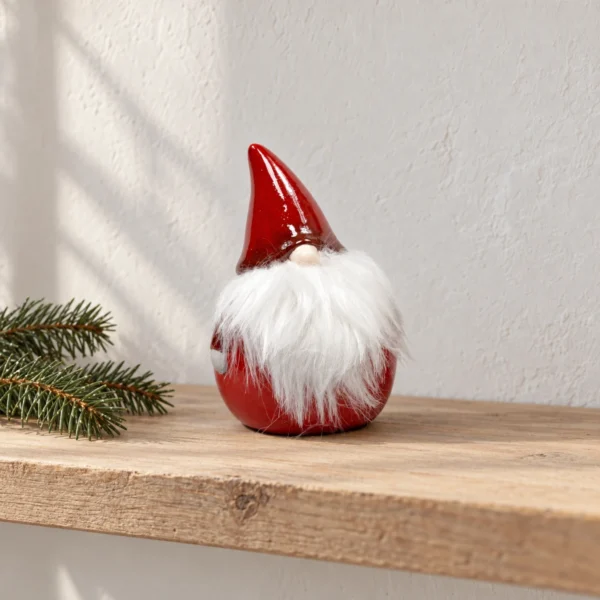 Röd tomte med vitt textilskägg stående på en ljus trähylla bredvid två grankvistar