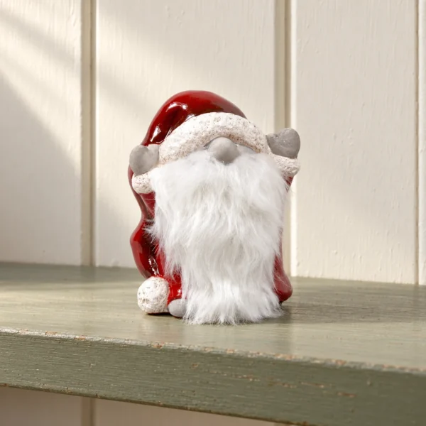 Tomte med röd luva, grå vantar och vitt skägg på en dovt grön trähylla.