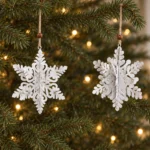 Två tredimensionella snöflingor i metall hänger på olika höjd i en julgran med varma ljuspunkter.