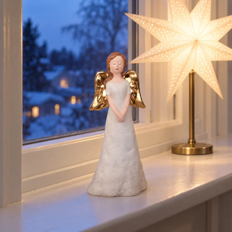 Ängel 27,5 cm i fönster med lysande julstjärna Ängel med vit glitterklänning och guldfärgade vingar på en vit fönsterbräda bredvid en lysande julstjärna på fot i vinterkväll.