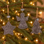 Stjärna, ängel och gran i mörk metall med XMAS-brickor hänger i snören runt julgranens grenar.