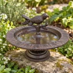 Mörkbrunt fågelbad i gjutjärn på en natursten bland grönska och vita blommor